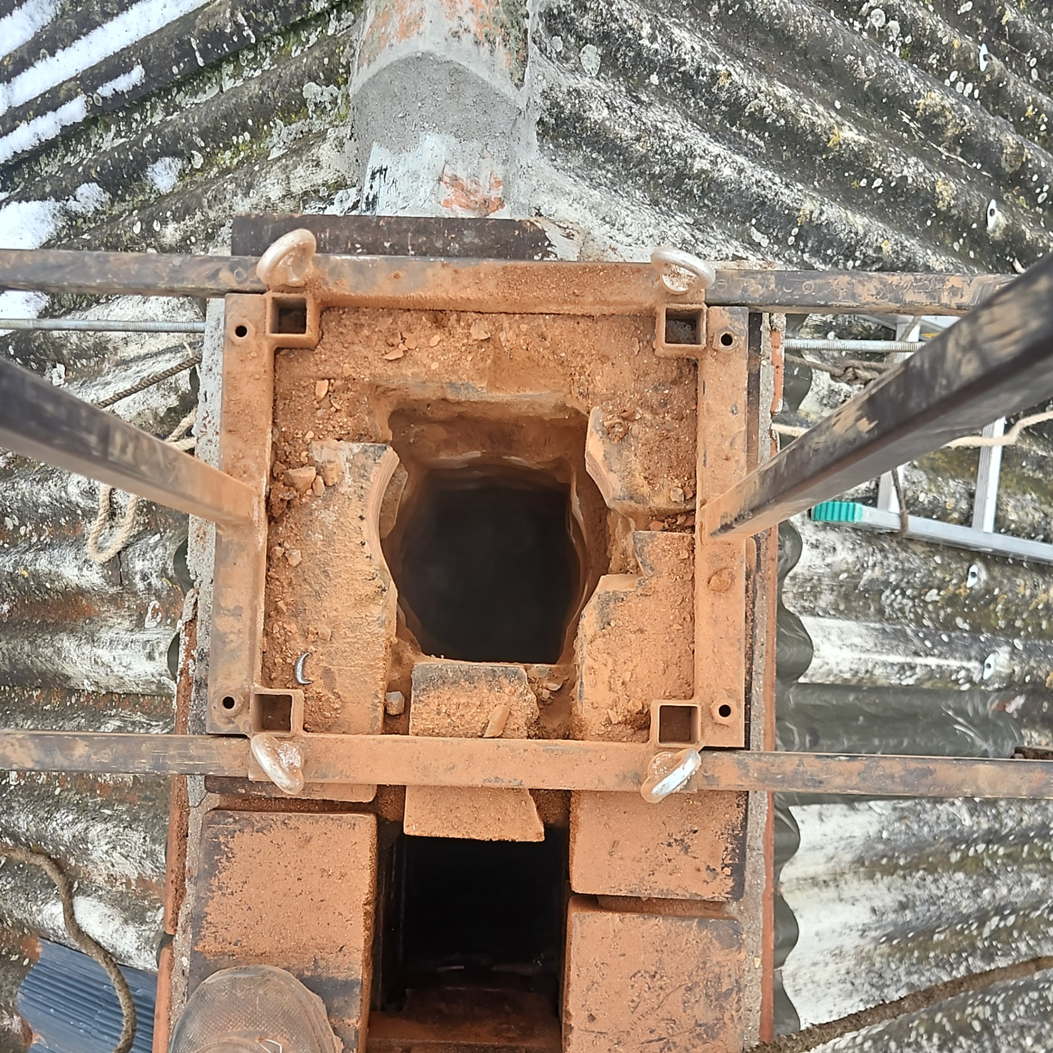 Otwarty komin z cegły, widok z góry, z metalową konstrukcją zabezpieczającą. Widoczny fragment dachu pokrytego eternitem i drabina kominiarska.