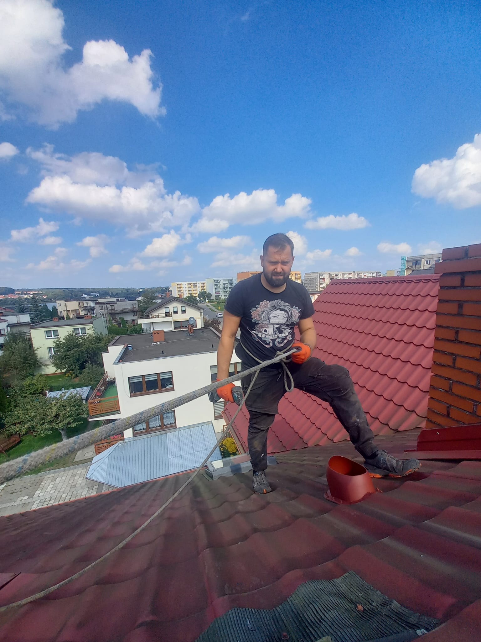 Pracownik w pomarańczowych rękawicach na dachu z czerwonej dachówki, zabezpieczony liną, montuje element komina. W tle zabudowa miejska i błękitne niebo.