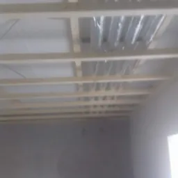 Stelaż drewniany pod sufitem z widoczną instalacją wentylacyjną w folii, szara ściana w tle.