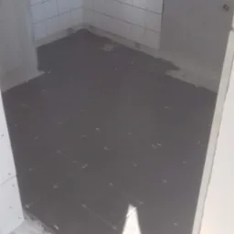 Układanie ciemnych płytek podłogowych w łazience, widoczne krzyżyki dystansowe, białe płytki na ścianach, surowy stan wykończenia.