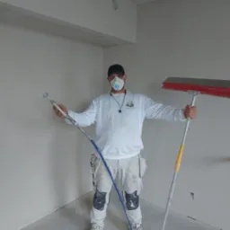 Pracownik w masce ochronnej, ubrudzony farbą, trzymający w dłoniach pistolet natryskowy do gładzi i długą szpachlę na teleskopowym kiju, w surowym wnętrzu z przygotowanymi do malowania ścianami.