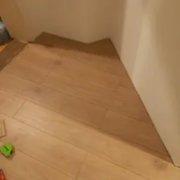 Ułożone panele podłogowe w rogu pokoju, przy białej ścianie, widoczna miara i fragment narzędzia.