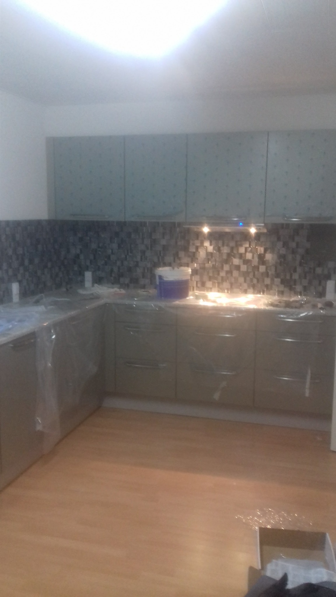 Kuchnia w trakcie prac wykończeniowych: zamontowane szafki z szarymi frontami, mozaika na ścianie nad blatem, podłoga wyłożona panelami, elementy zabezpieczone folią, widoczne wiadro z farbą.