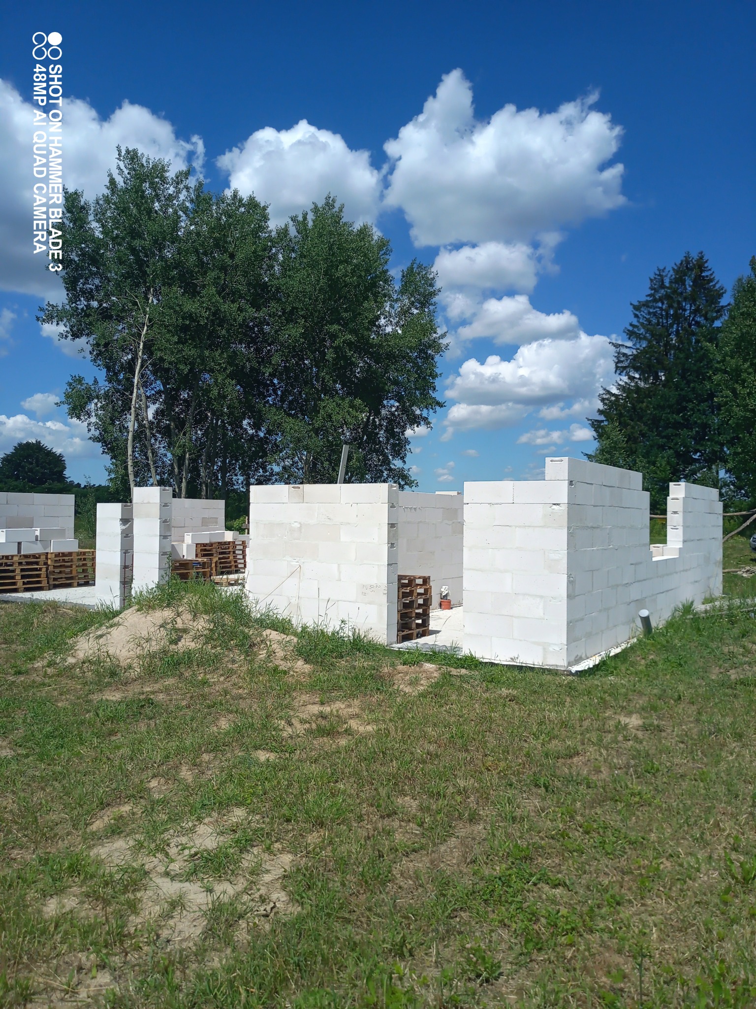 Wznoszone mury domu z bloczków betonu komórkowego na tle błękitnego nieba z białymi chmurami, palety z bloczkami obok, widok z poziomu trawy.