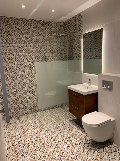 Nowoczesna łazienka z wzorzystymi płytkami w odcieniach beżu i brązu na podłodze i ścianach, kabina prysznicowa ze szkła, wisząca toaleta i umywalka z szafką z ciemnego drewna.