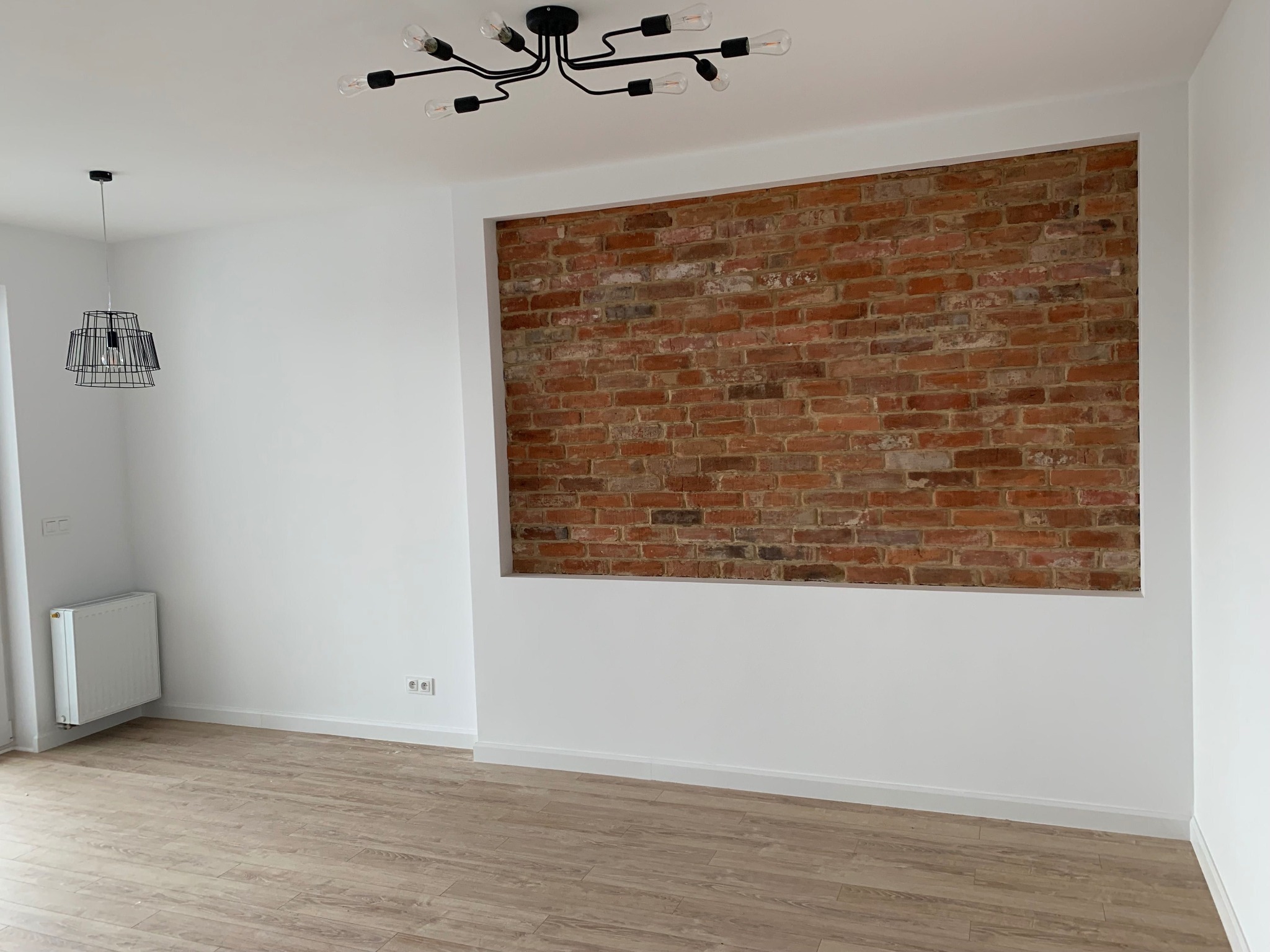 Pusty pokój po remoncie z drewnopodobną podłogą, białe ściany i fragment ściany z czerwonej cegły w prostokątnej wnęce, czarny żyrandol sufitowy i wisząca lampa z drucianym kloszem.