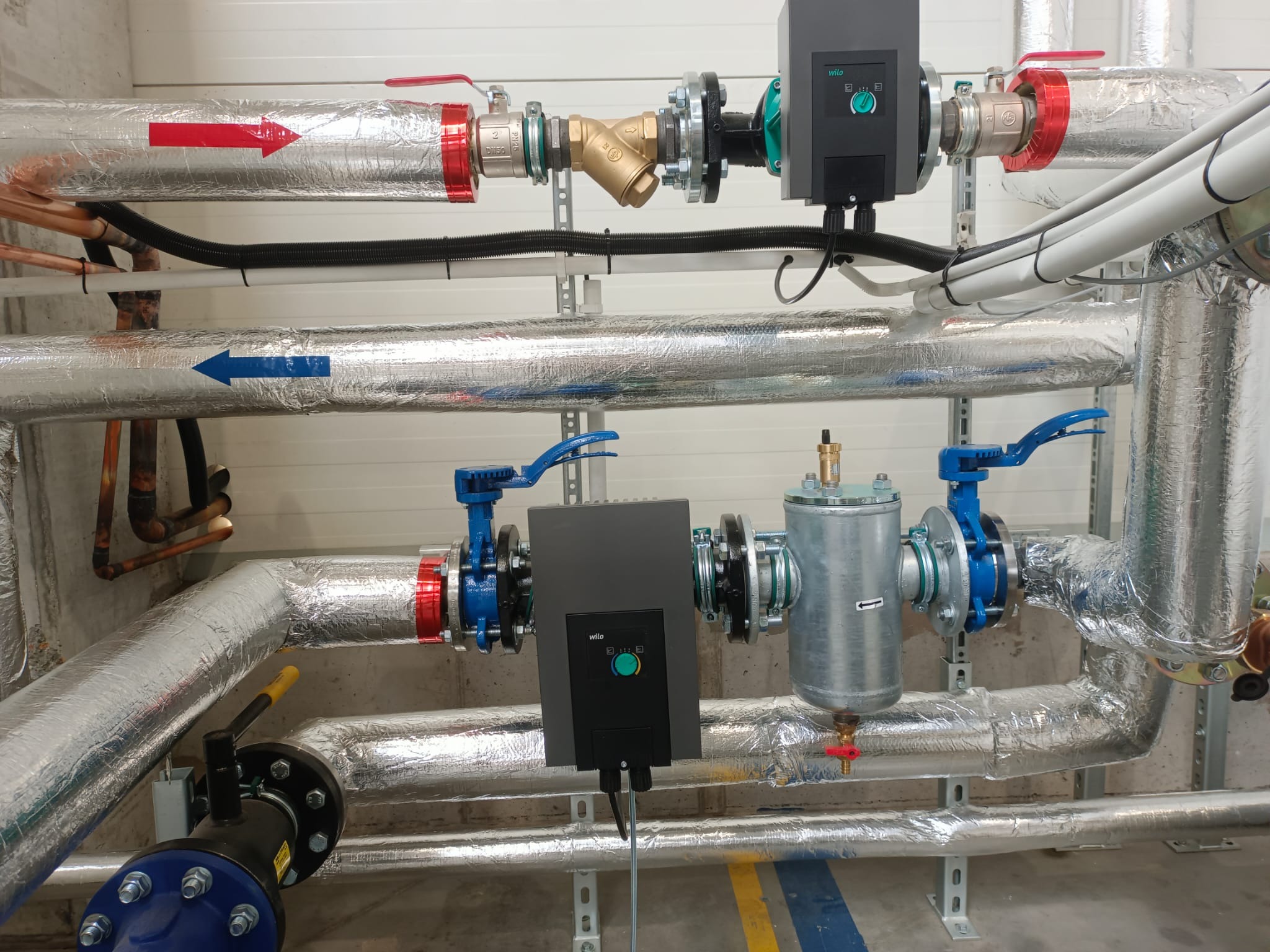 Kotłownia z rurami pokrytymi folią izolacyjną, zaworami z niebieskimi uchwytami i czarnymi elementami sterującymi Wilo, widoczne strzałki wskazujące kierunek przepływu na rurach.