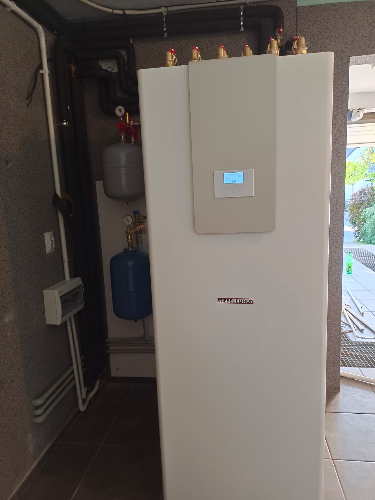 Kotłownia z pompą ciepła Stiebel Eltron typu monoblok powietrze-woda, widoczne rury, zbiornik buforowy i zasobnik CWU, zainstalowane przy szarej ścianie w pomieszczeniu technicznym.