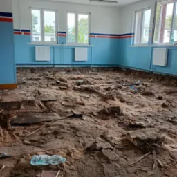 Wnętrze pomieszczenia w trakcie rozbiórki podłogi, widoczne resztki gruzu, cegieł i drewnianych elementów na ziemi, ściany pomalowane na niebiesko z czerwonym paskiem, okna otwarte, butelka wody...