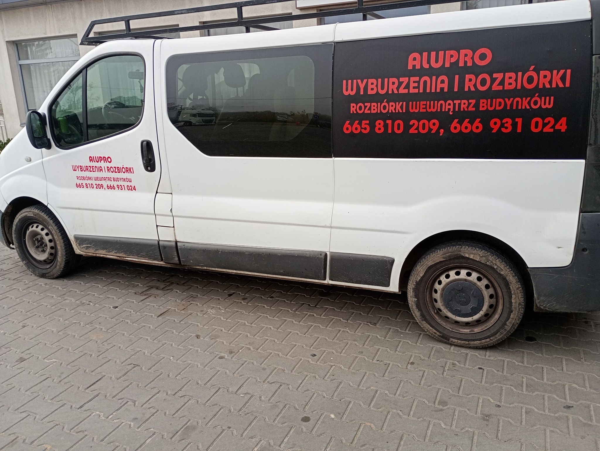 Biały samochód dostawczy z czerwonym napisem 'ALUPRO Wyburzenia i Rozbiórki Rozbiórki wewnątrz budynków' oraz numerami telefonów, zaparkowany na chodniku z kostki brukowej.