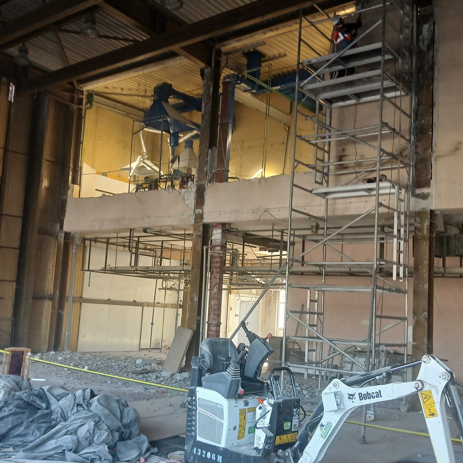 Wnętrze budynku w trakcie rozbiórki z widocznym minikoparką Bobcat, rusztowaniem i częściowo usuniętą ścianą. Gruz i zabezpieczenia na podłodze.