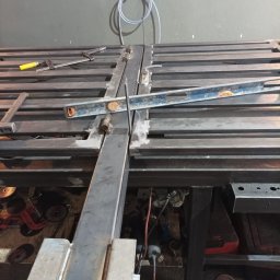 CraftsMan - Metalowa konstrukcja spawana na stole warsztatowym, poziomica i narzędzia w tle, widoczne ślady spawania.