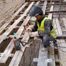 CraftsMan - Pracownik w hełmie spawalniczym i kamizelce odblaskowej, kuca pomiędzy stalowymi belkami, spawarka obok, w tle ceglana ściana z oknem.