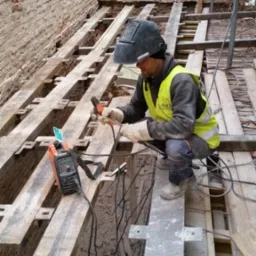 Pracownik w hełmie spawalniczym i kamizelce odblaskowej, kuca pomiędzy stalowymi belkami, spawarka obok, w tle ceglana ściana z oknem.