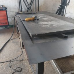 CraftsMan - Żółta szlifierka kątowa leży na metalowym blacie warsztatowym, w tle spawarka i metalowe konstrukcje.