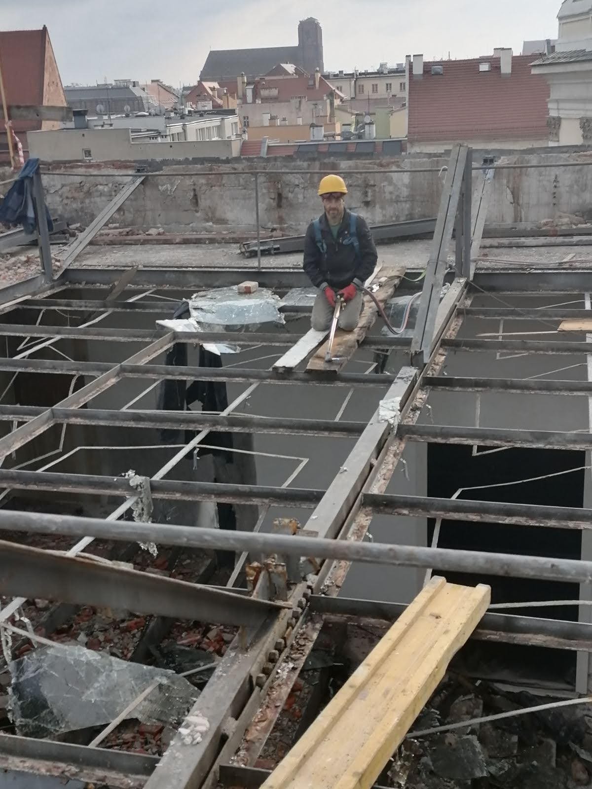 Pracownik w kasku i rękawicach klęczy na metalowej konstrukcji dachu podczas demontażu, w tle budynki miejskie i kościół.