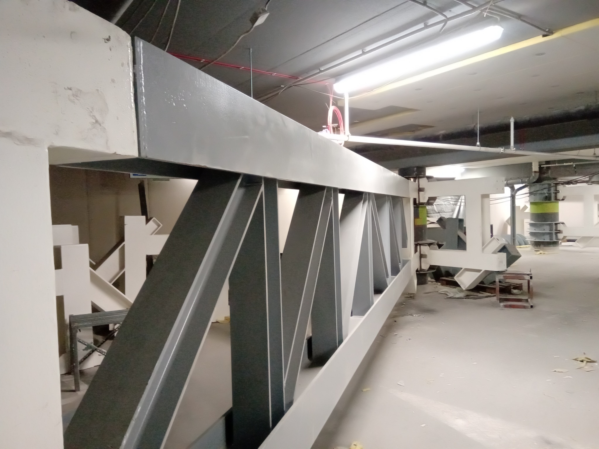 Wnętrze hali w trakcie budowy, widoczne elementy konstrukcji stalowej pomalowane na szaro i biało, sufit z instalacjami i oświetleniem, na podłodze widoczne ślady budowy.