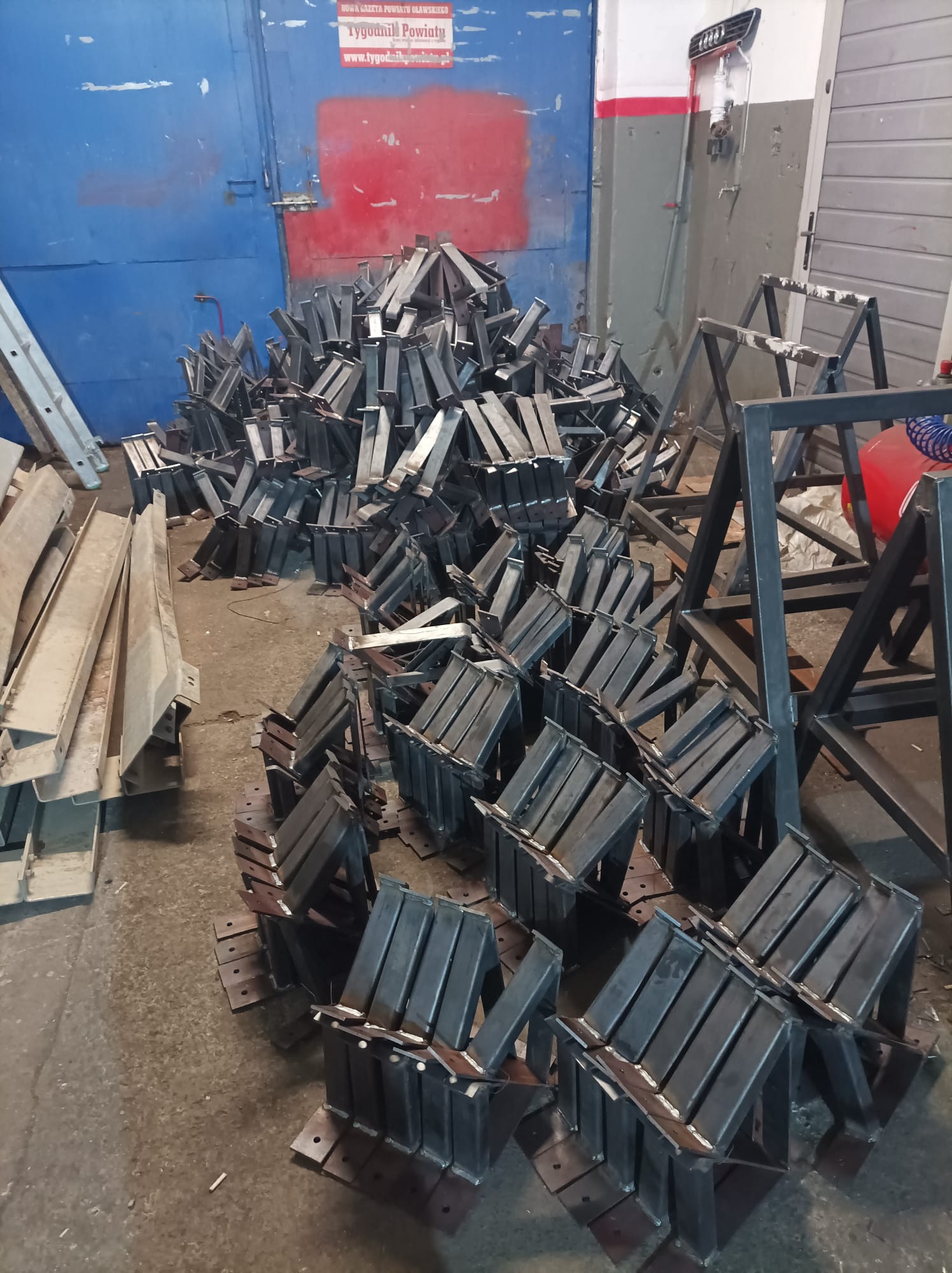 Stalowe elementy konstrukcyjne, prawdopodobnie podpory lub wsporniki, ułożone w stos w hali produkcyjnej, widoczna niebieska brama i fragment drabiny.
