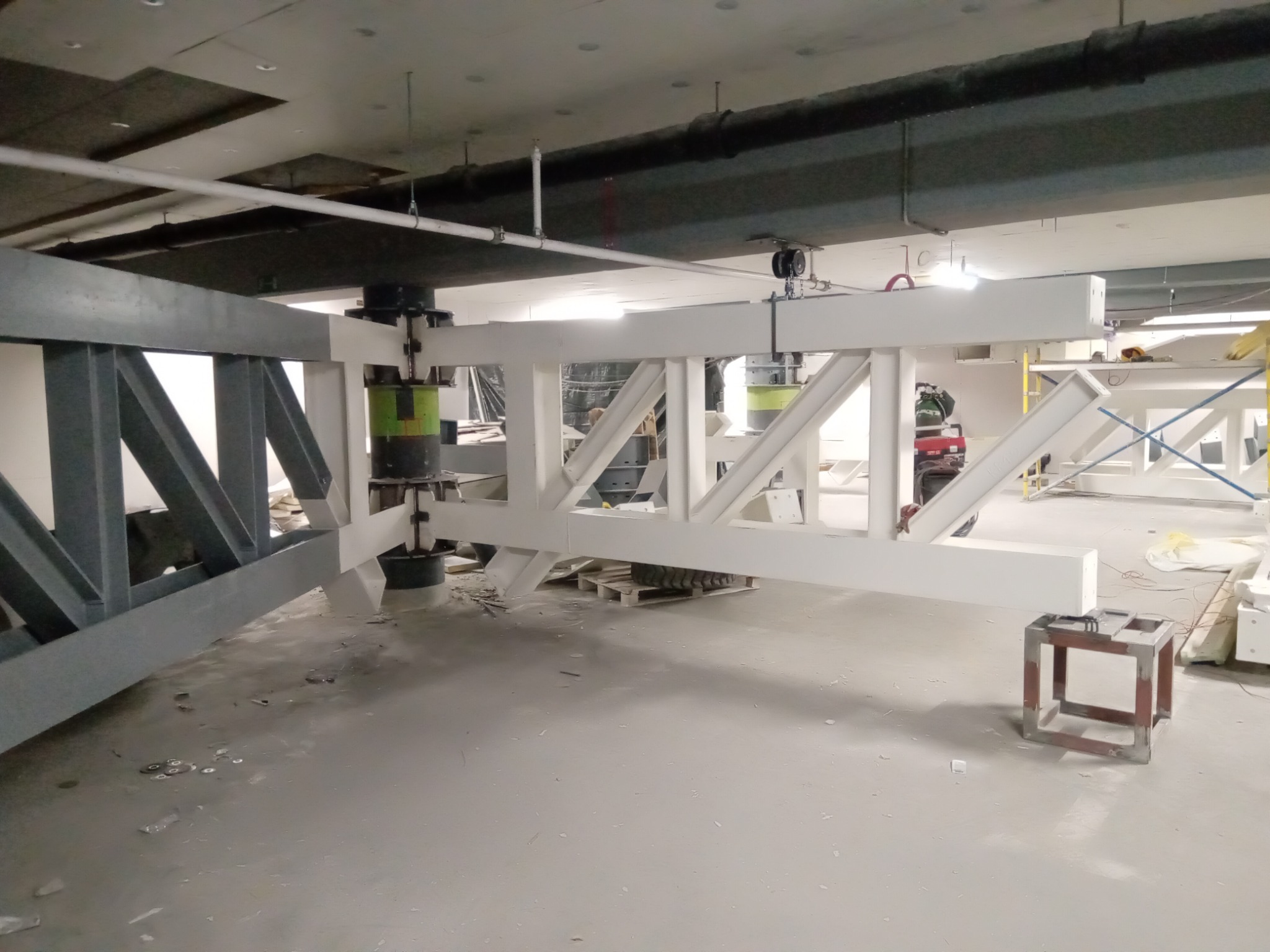 Wnętrze hali w trakcie budowy, widoczne stalowe elementy konstrukcyjne w kolorze szarym i białym, podtrzymywane przez zielone cylindry, sufit z instalacjami.