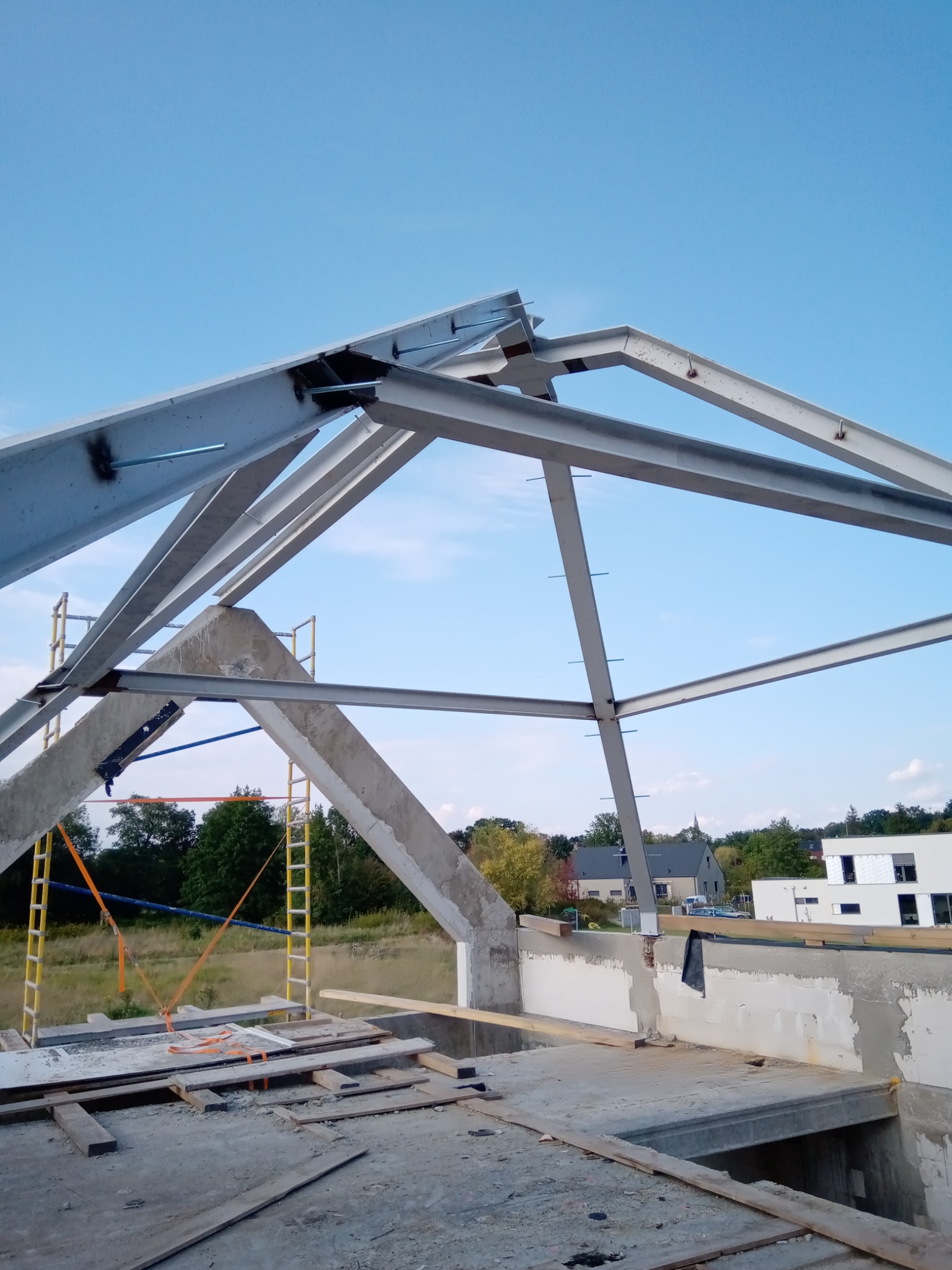 Konstrukcja stalowa dachu hali w trakcie budowy, widoczne elementy zbrojenia i rusztowania na tle błękitnego nieba i zabudowań.