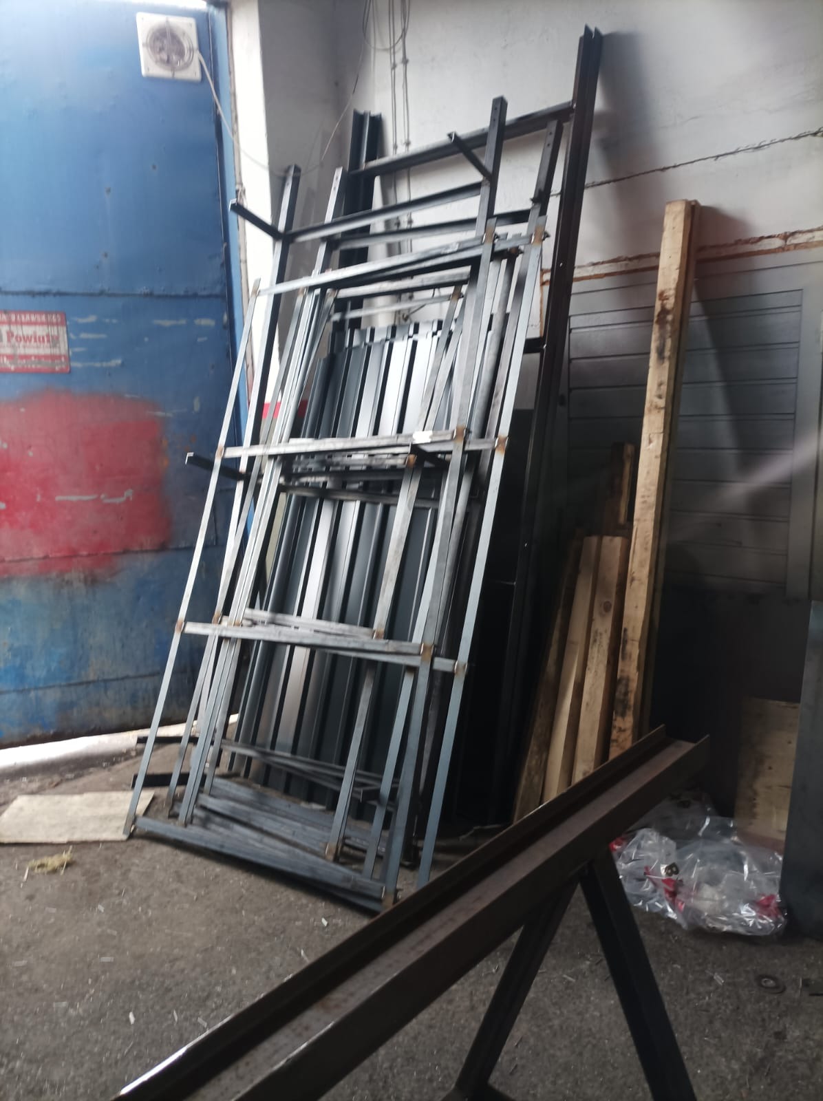 Stalowe ramy konstrukcyjne o regularnych kształtach, oparte o ścianę w warsztacie, obok deski i metalowe profile.