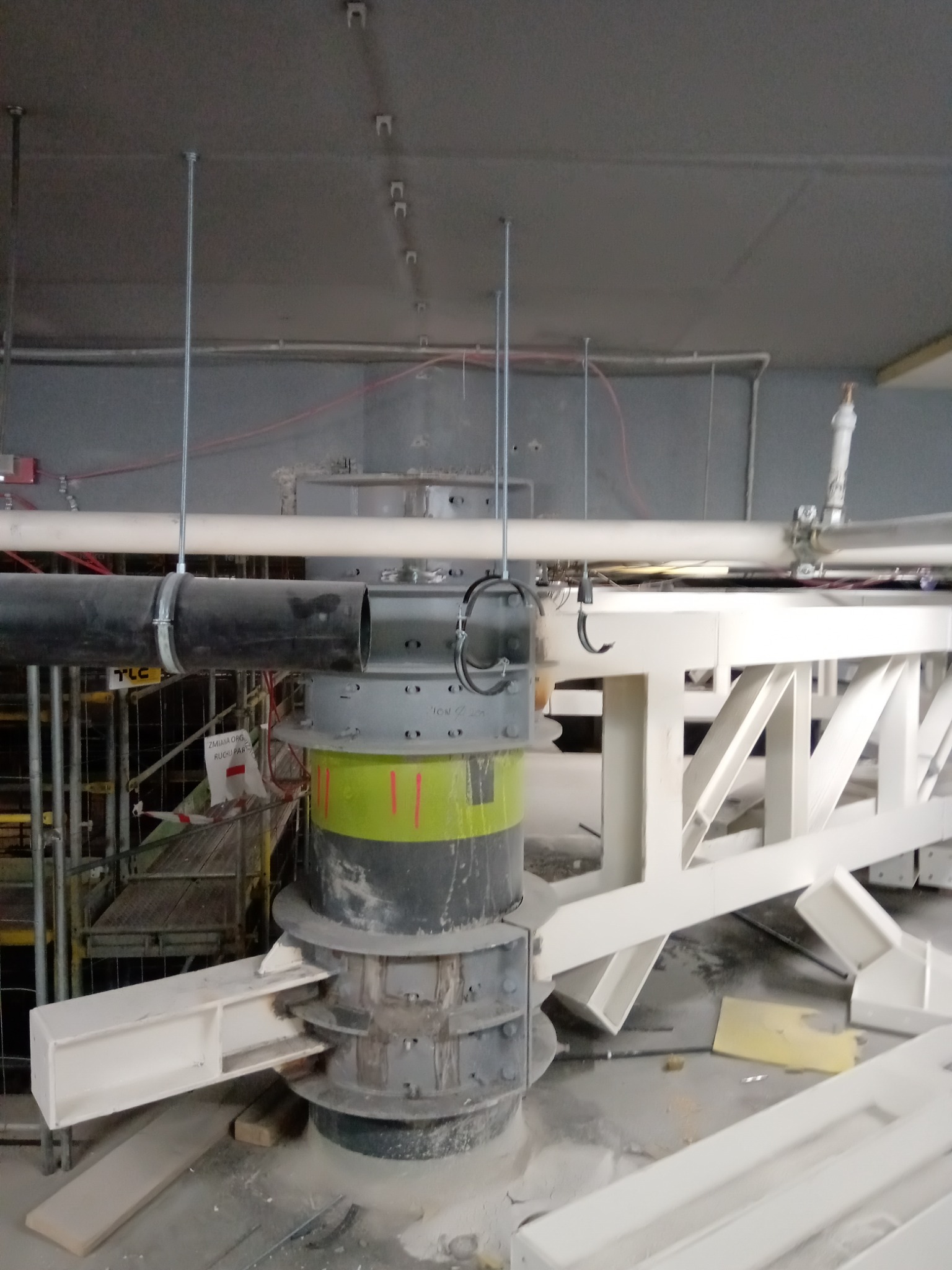 Wnętrze hali w trakcie budowy, widoczna stalowa konstrukcja wsporcza, rury instalacyjne i fragment rusztowania.