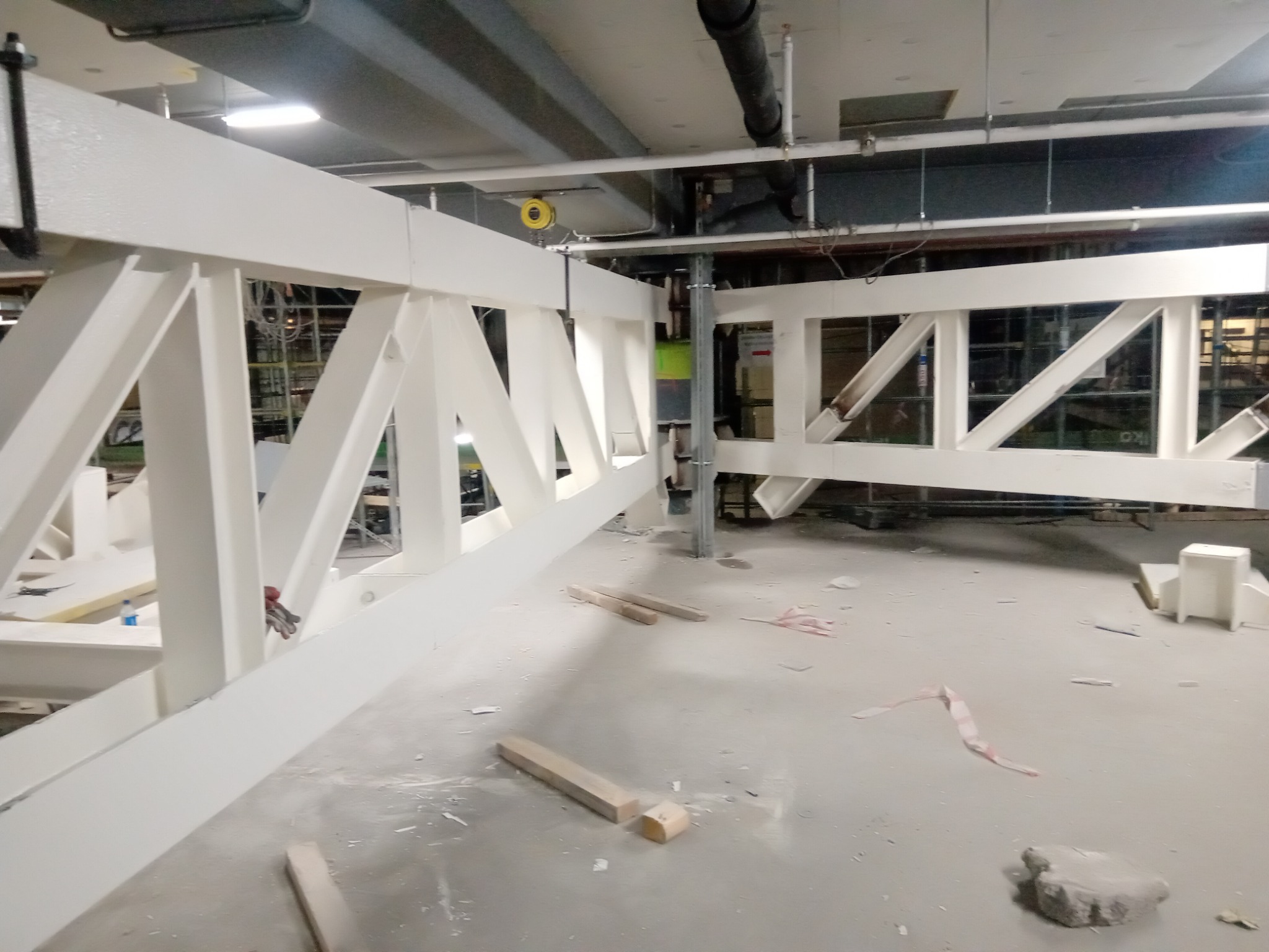 Białe, stalowe konstrukcje kratowe w hali produkcyjnej, widoczne elementy montażowe i fragmenty podłogi z drobnymi odpadami.