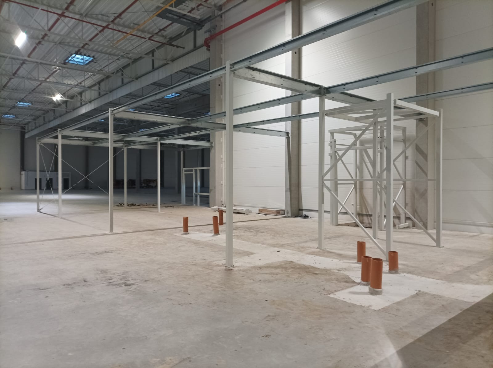 Wnętrze hali w trakcie budowy z widoczną stalową konstrukcją szkieletową i pionowymi rurami kanalizacyjnymi wystającymi z posadzki.