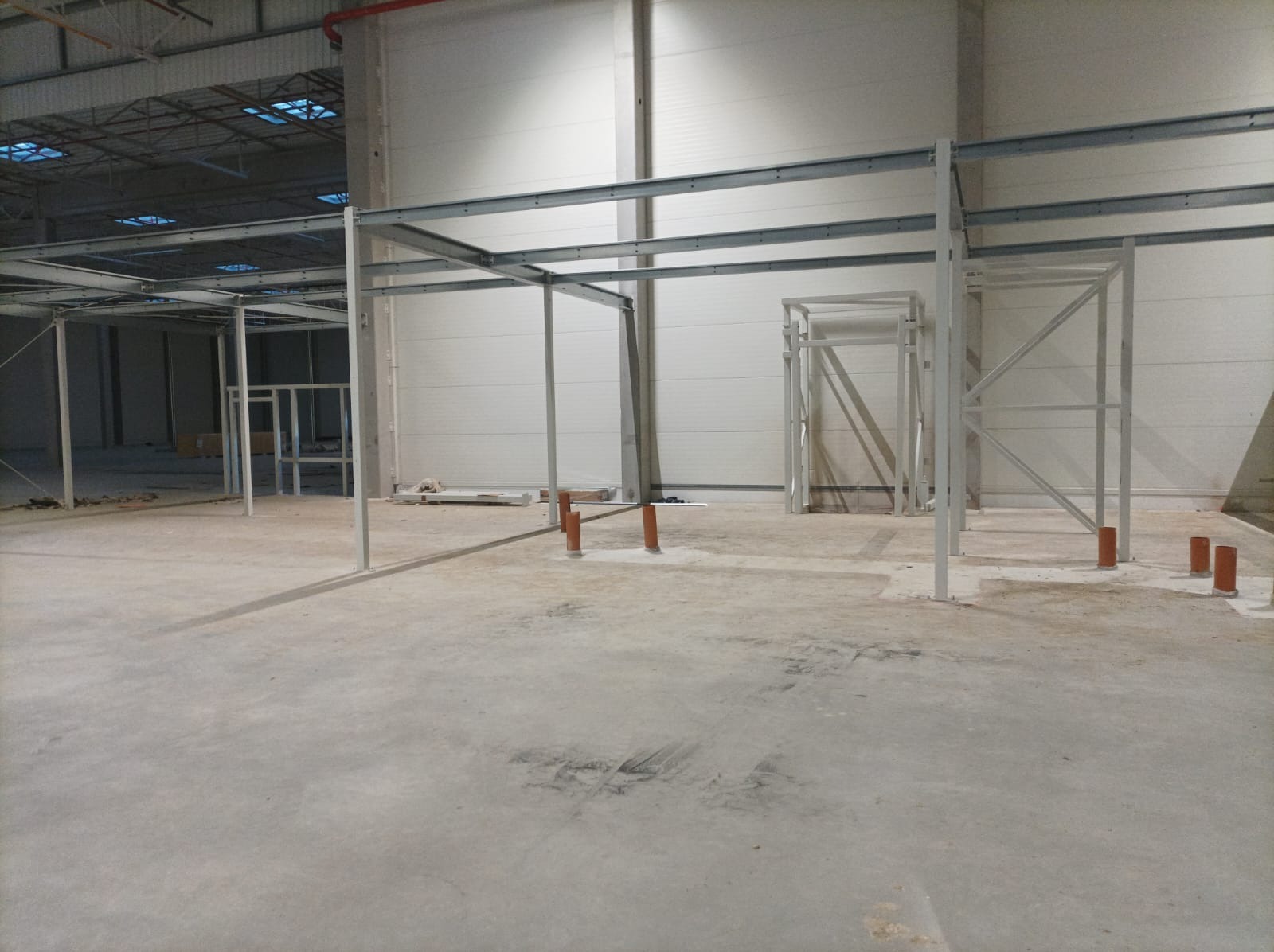 Wnętrze hali w trakcie budowy, widoczne stalowe konstrukcje szkieletowe, betonowa posadzka z plamami i rury kanalizacyjne wystające z podłoża.