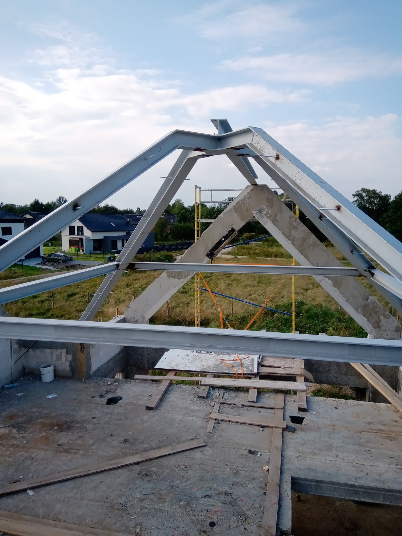 Wnętrze budowy hali z widoczną stalową konstrukcją dachu, częściowo zabetonowaną, na tle krajobrazu z domami i zielenią.