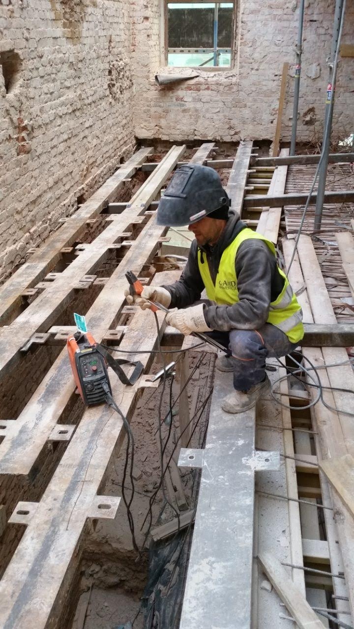 Pracownik w hełmie spawalniczym i kamizelce odblaskowej, kuca pomiędzy stalowymi belkami, spawarka obok, w tle ceglana ściana z oknem.