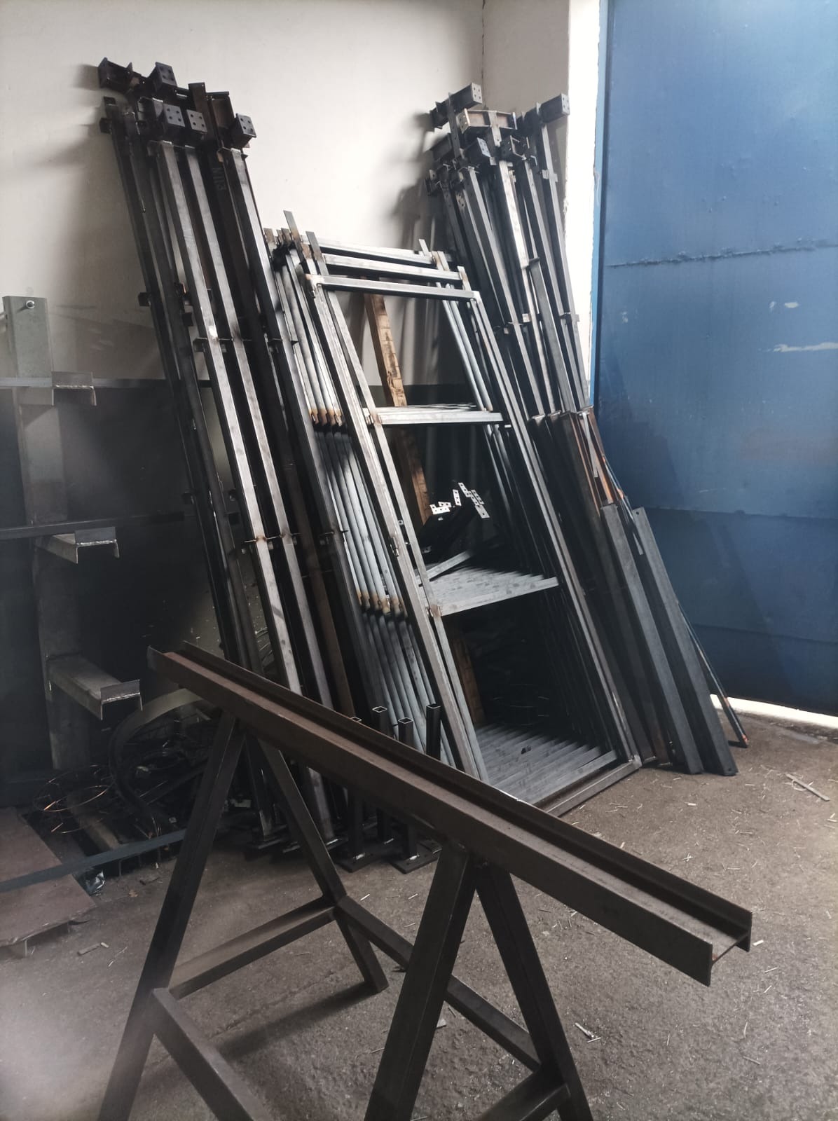 Stalowe elementy konstrukcyjne, profile i stojaki składowane w warsztacie, widoczne ślady obróbki i spawania.