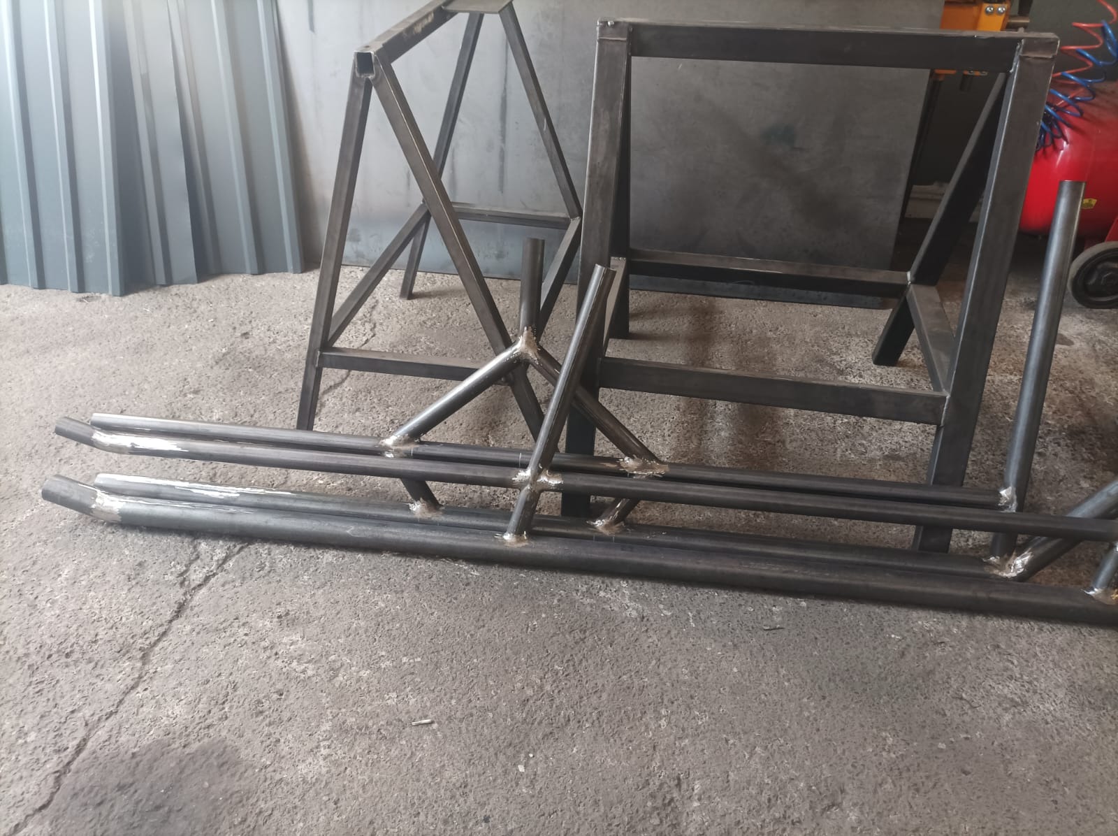 Surowe, spawane konstrukcje stalowe: dwie ramy w kształcie litery A i prostokątna, oraz elementy rurowe z widocznymi spawami, leżące na betonowej podłodze warsztatu.