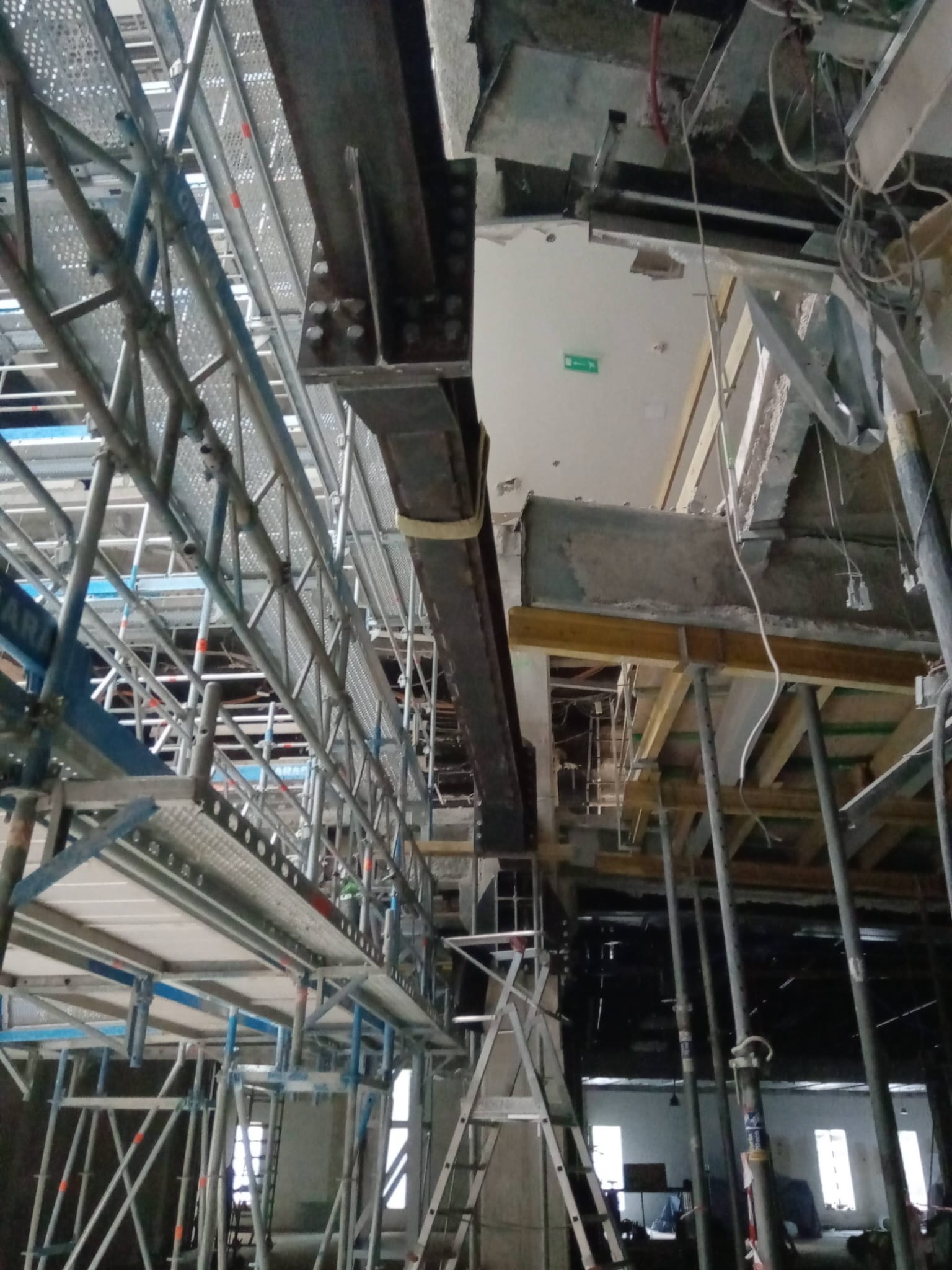 Wnętrze budynku w trakcie budowy z widoczną konstrukcją stalową, rusztowaniami i elementami betonowymi, oświetlenie mieszane.