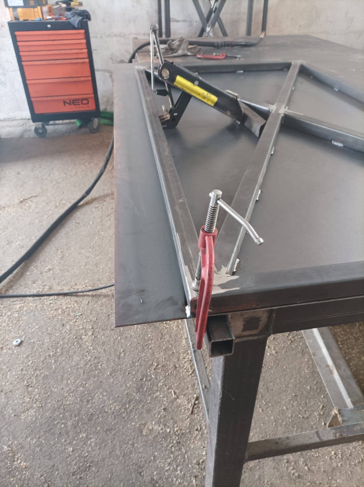 Metalowa konstrukcja ramy spawana na stole warsztatowym, zabezpieczona ściskami, z widocznym pomarańczowym wózkiem narzędziowym NEO w tle.