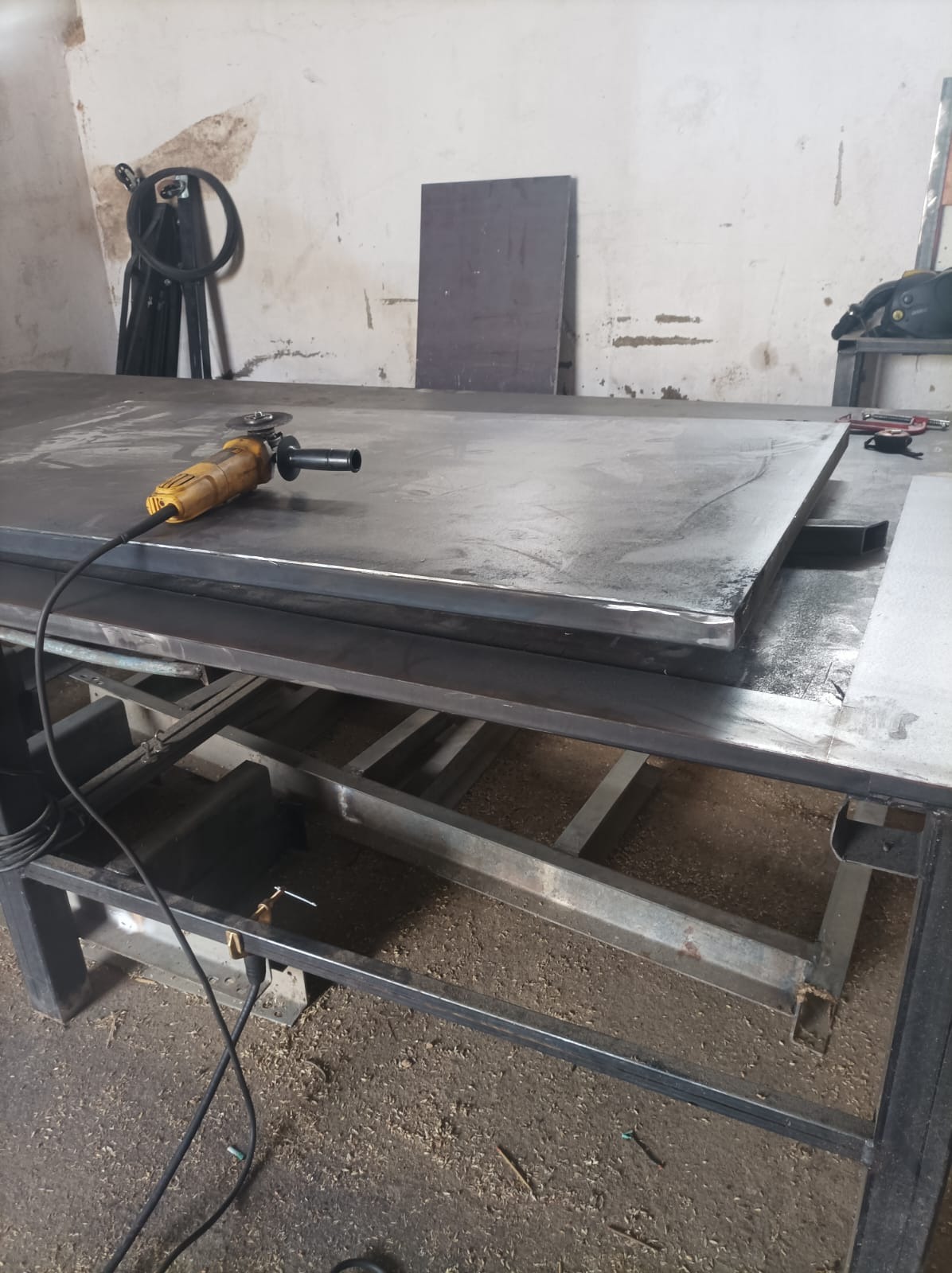 Żółta szlifierka kątowa leży na dużej, metalowej płycie na metalowym stole warsztatowym w warsztacie, w tle widoczna ściana z plamami i metalowe elementy.