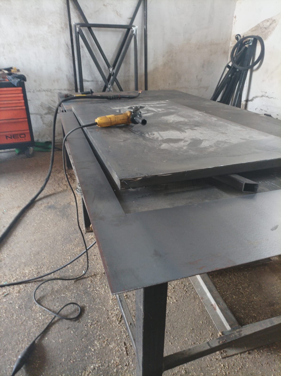 Żółta szlifierka kątowa leży na metalowym blacie warsztatowym, w tle spawarka i metalowe konstrukcje.