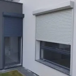 Dwa okna z zamontowanymi roletami zewnętrznymi: szara roleta po prawej stronie i grafitowa po lewej, na tle jasnej ściany budynku.