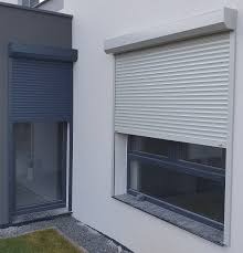 Dwa okna z zamontowanymi roletami zewnętrznymi: szara roleta po prawej stronie i grafitowa po lewej, na tle jasnej ściany budynku.