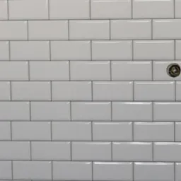 Ściana wyłożona błyszczącymi, białymi płytkami typu metro z ciemnoszarą fugą; widoczne gniazdko elektryczne bez osłonki.