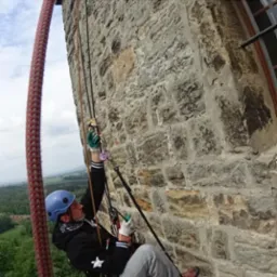 Pracownik w kasku i rękawicach, zawieszony na linach przy kamiennej ścianie zamku, wykonuje prace na wysokości z użyciem specjalistycznego sprzętu alpinistycznego.