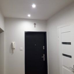 JANKOS PIOTR JANKOWSKI - Korytarz po odświeżeniu ścian, z dwiema parami drzwi: czarne drzwi wejściowe pod krzyżem na ścianie i białe drzwi wewnętrzne z poziomymi wstawkami, oświetlenie punktowe sufitowe.