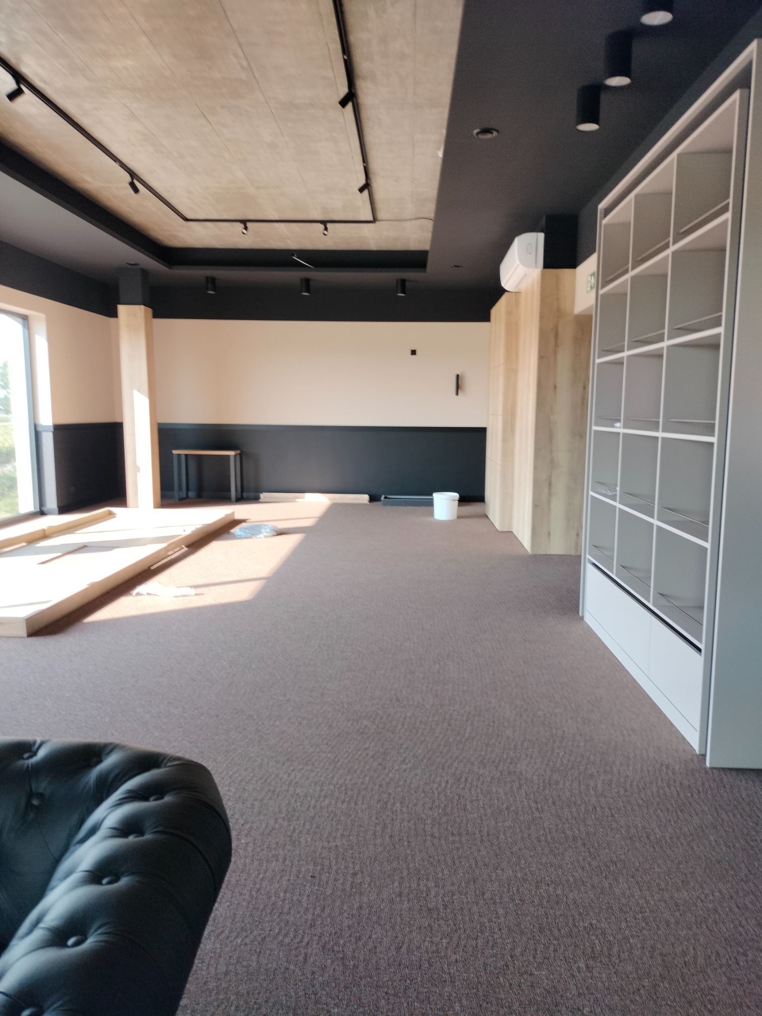 Wnętrze pomieszczenia w trakcie prac wykończeniowych, widoczne elementy konstrukcyjne, surowy betonowy sufit z szynoprzewodami i reflektorami, fragment sofy w rogu, częściowo zabudowana szafa...