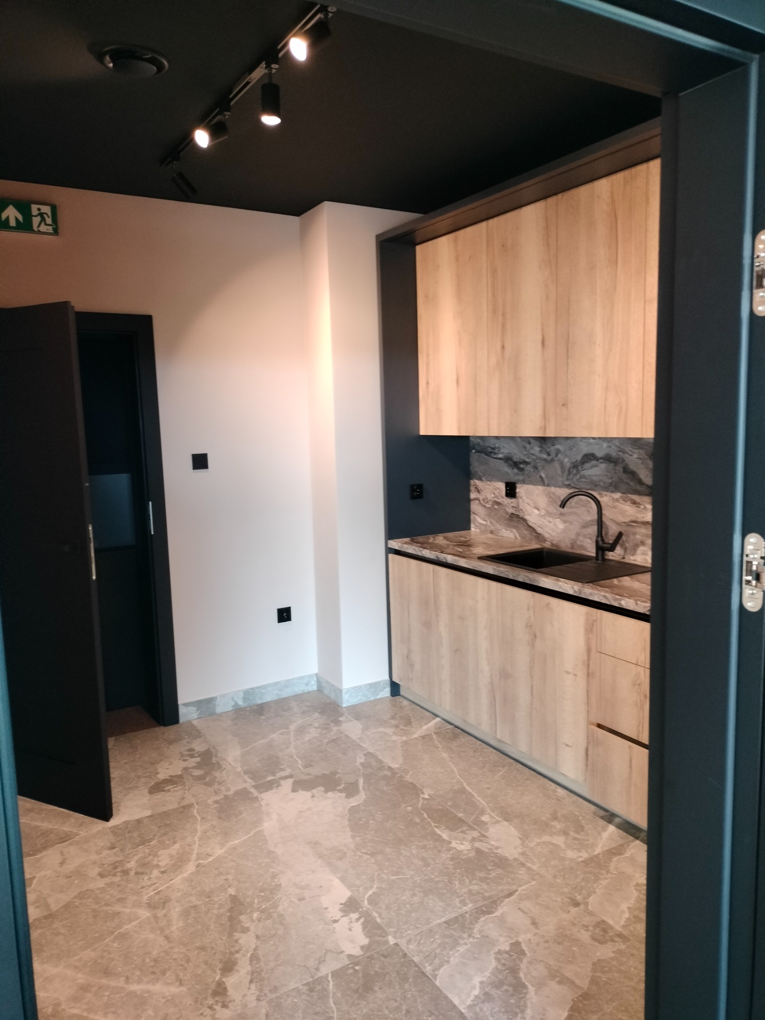 Nowoczesna kuchnia z drewnianymi szafkami, marmurowym blatem i czarną armaturą, widok z korytarza z podświetlanym sufitem i marmurową podłogą.