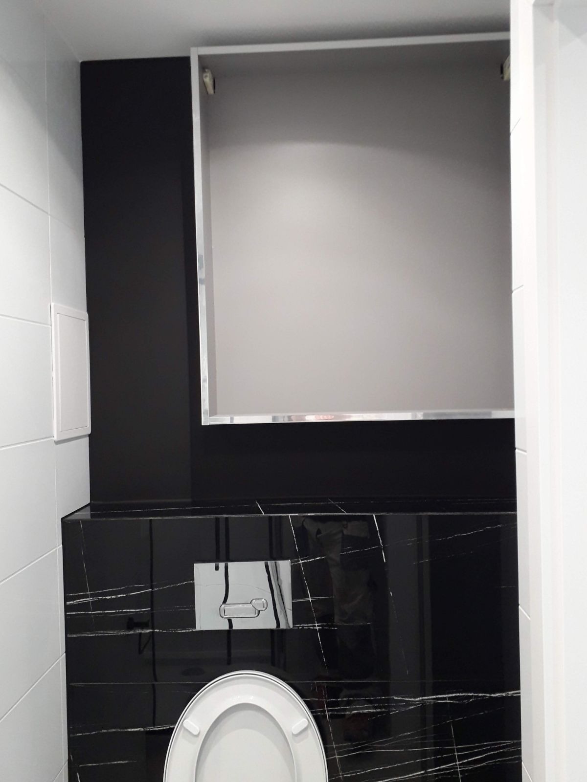 Nowoczesna toaleta z czarnymi płytkami imitującymi marmur, białymi płytkami na ścianie, ukryta szafka nad stelażem WC, chromowany przycisk spłukujący.