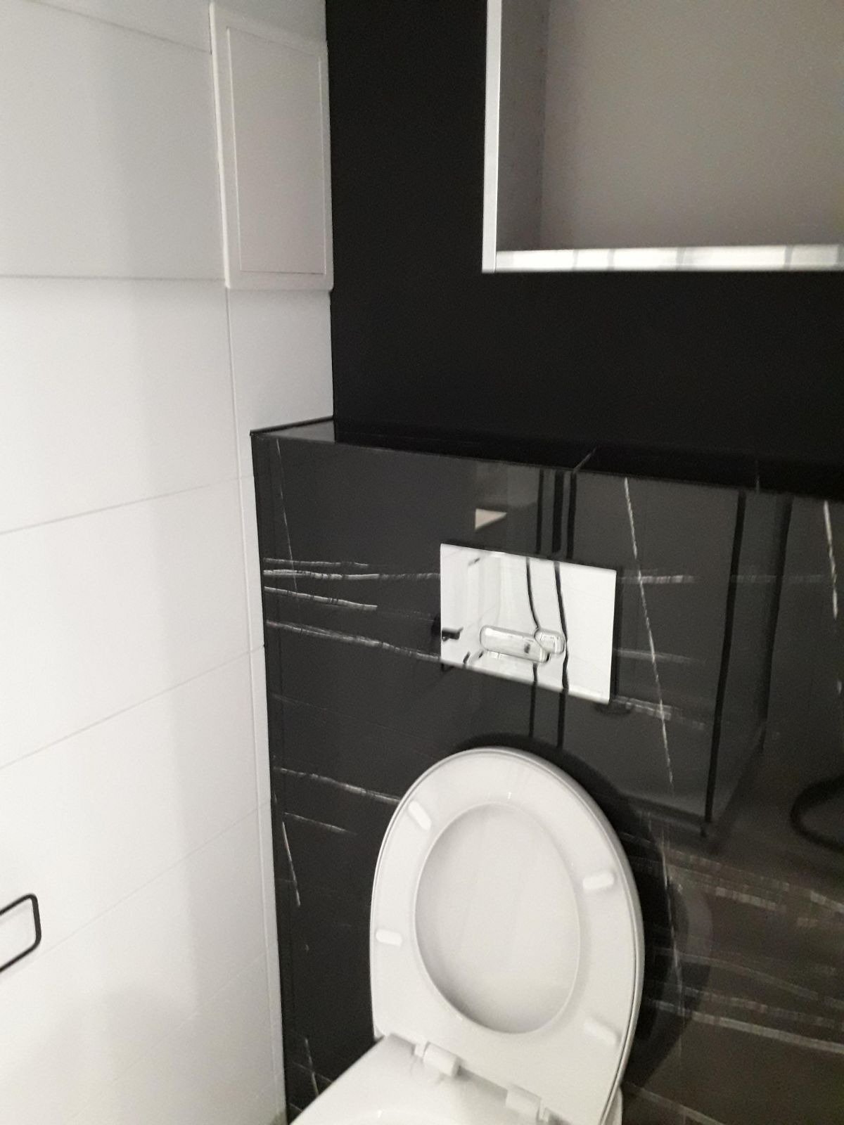 Nowoczesna toaleta z czarnymi marmurowymi płytkami i białymi płytkami ściennymi, widoczny chromowany przycisk spłukiwania i biała szafka wisząca.