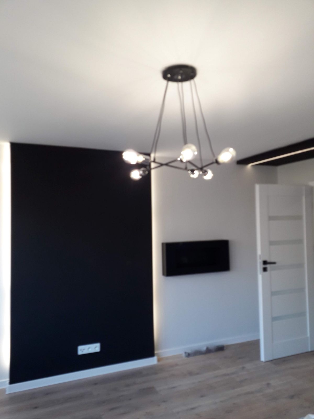 Nowoczesne wnętrze: czarna ściana z podświetleniem LED, designerska lampa wisząca z żarówkami Edison, białe drzwi z poziomymi panelami i drewnopodobna podłoga.