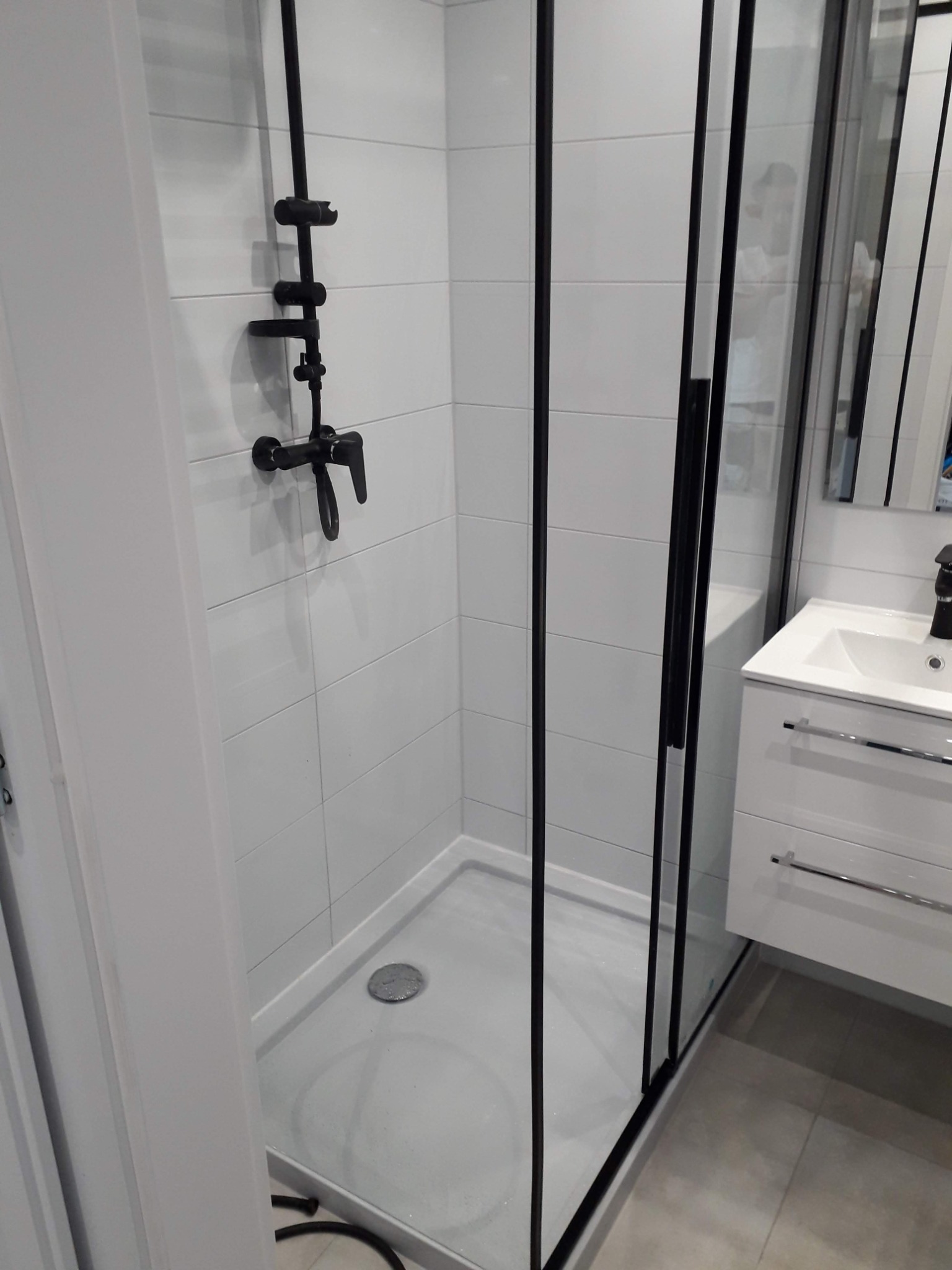 Nowoczesna kabina prysznicowa z czarną armaturą i białymi płytkami, widoczna umywalka z szafką w tle.