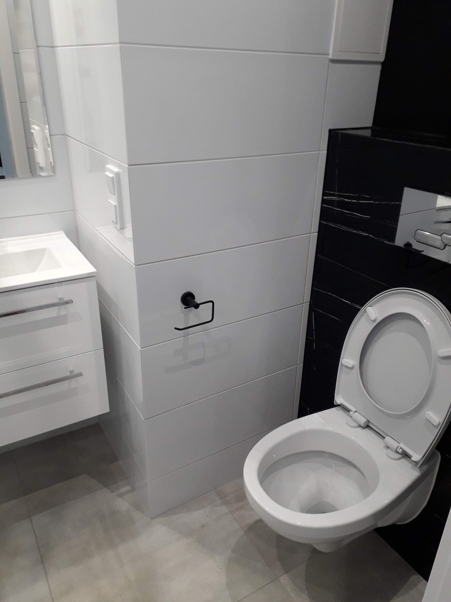 Nowoczesna łazienka z białymi, błyszczącymi płytkami ułożonymi horyzontalnie, wisząca toaleta z otwartą klapą, biała szafka z umywalką oraz czarny panel dekoracyjny z białymi żyłkami.