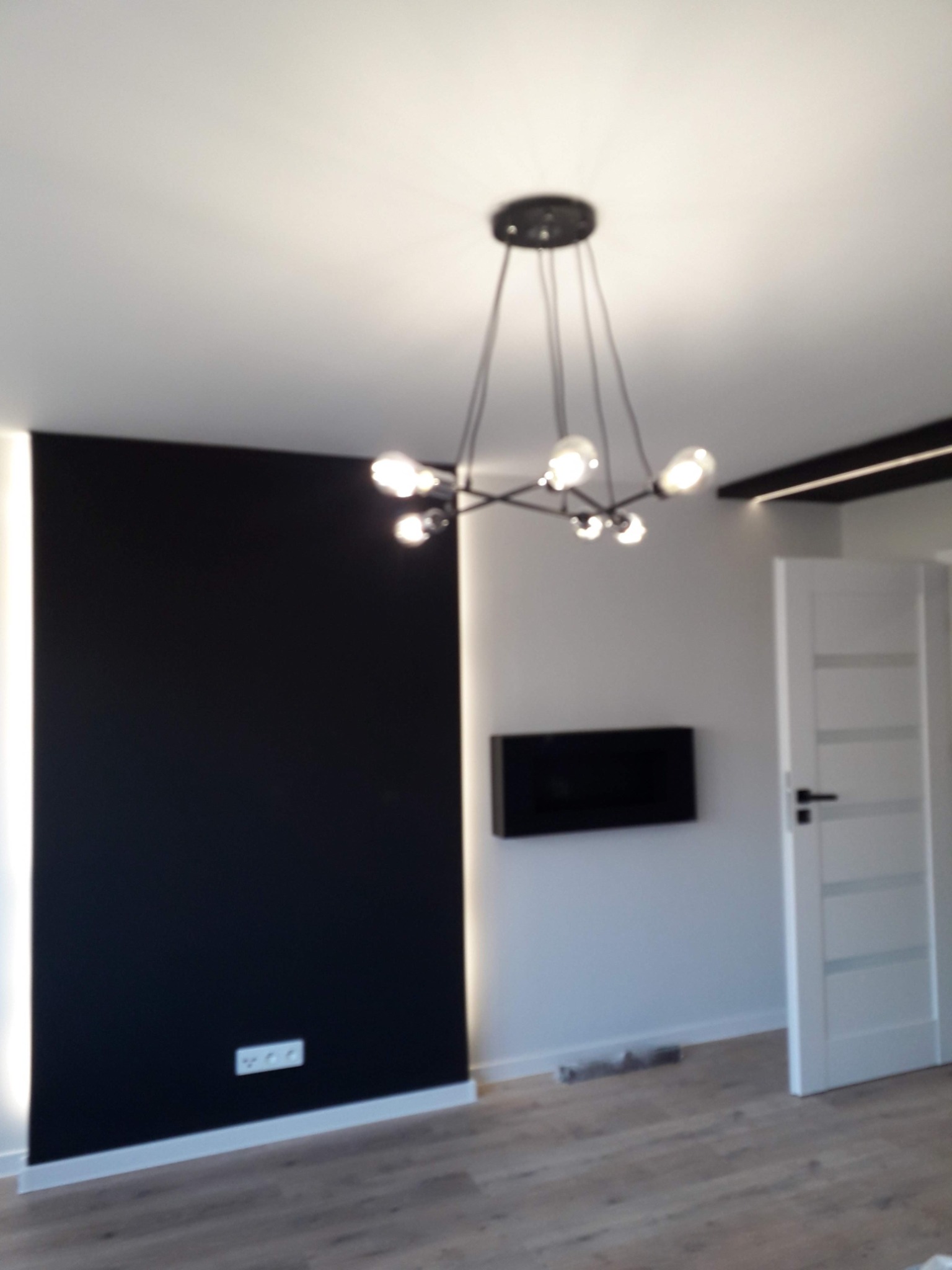 Nowoczesne wnętrze: Czarna ściana z oświetleniem LED, designerska lampa sufitowa z kulistymi żarówkami, białe drzwi z przeszkleniami i drewniana podłoga.